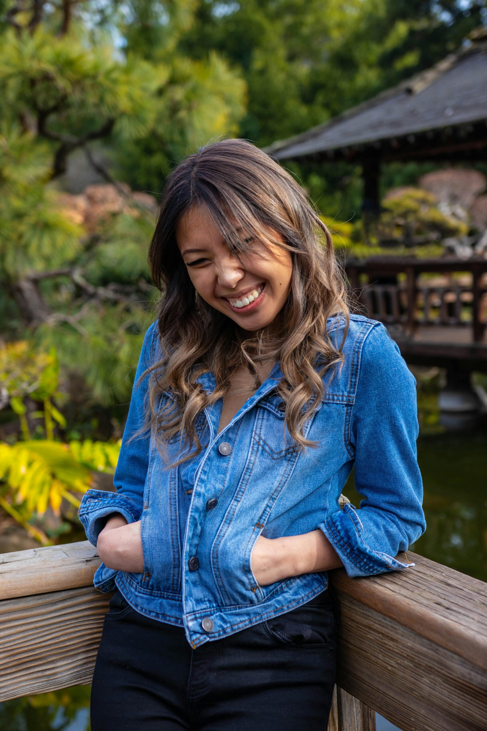 A woman in a denim jacket smiles joyfully by a wooden railing outdoors.