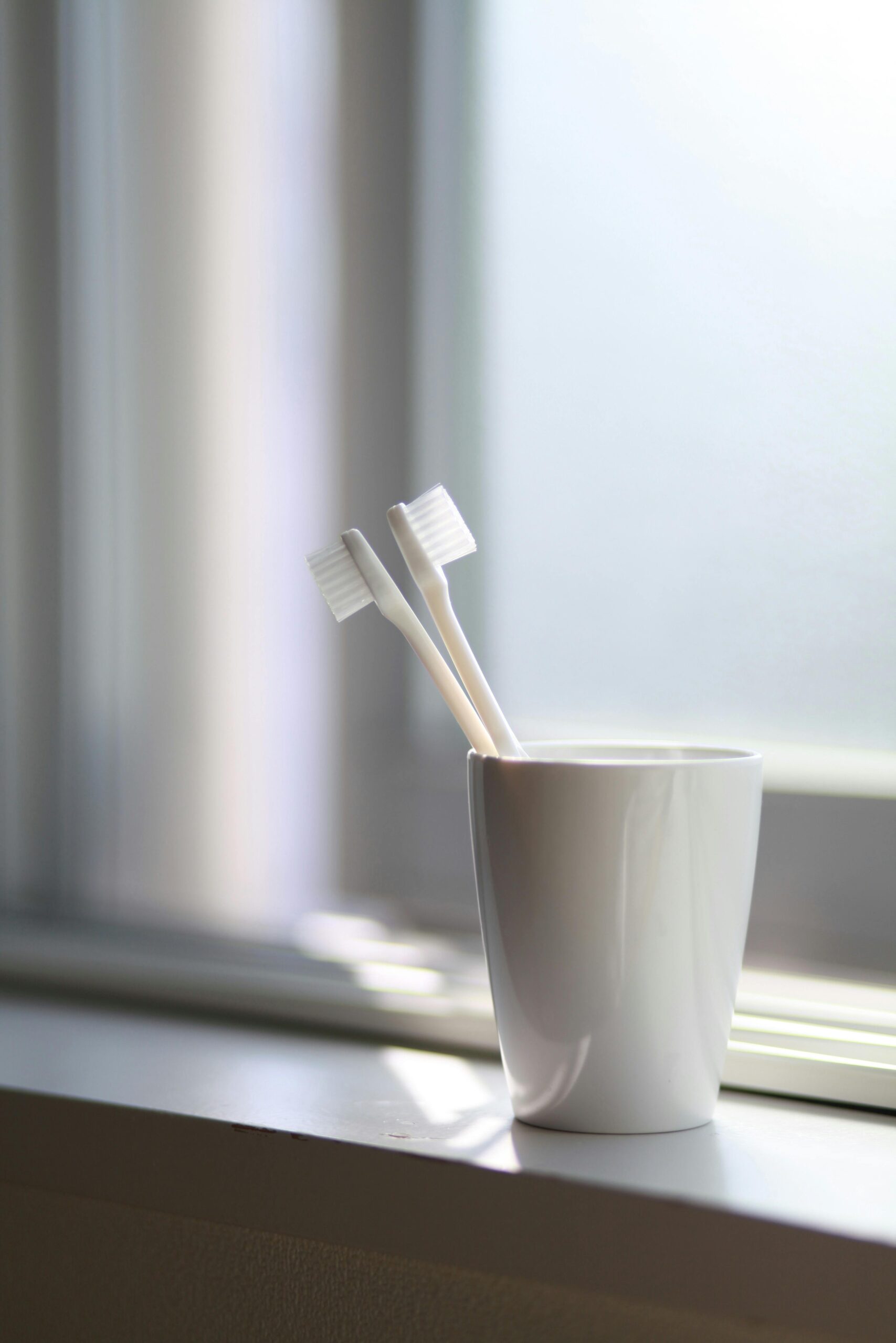 A simple, serene image of two toothbrushes in a white cup on a windowsill.