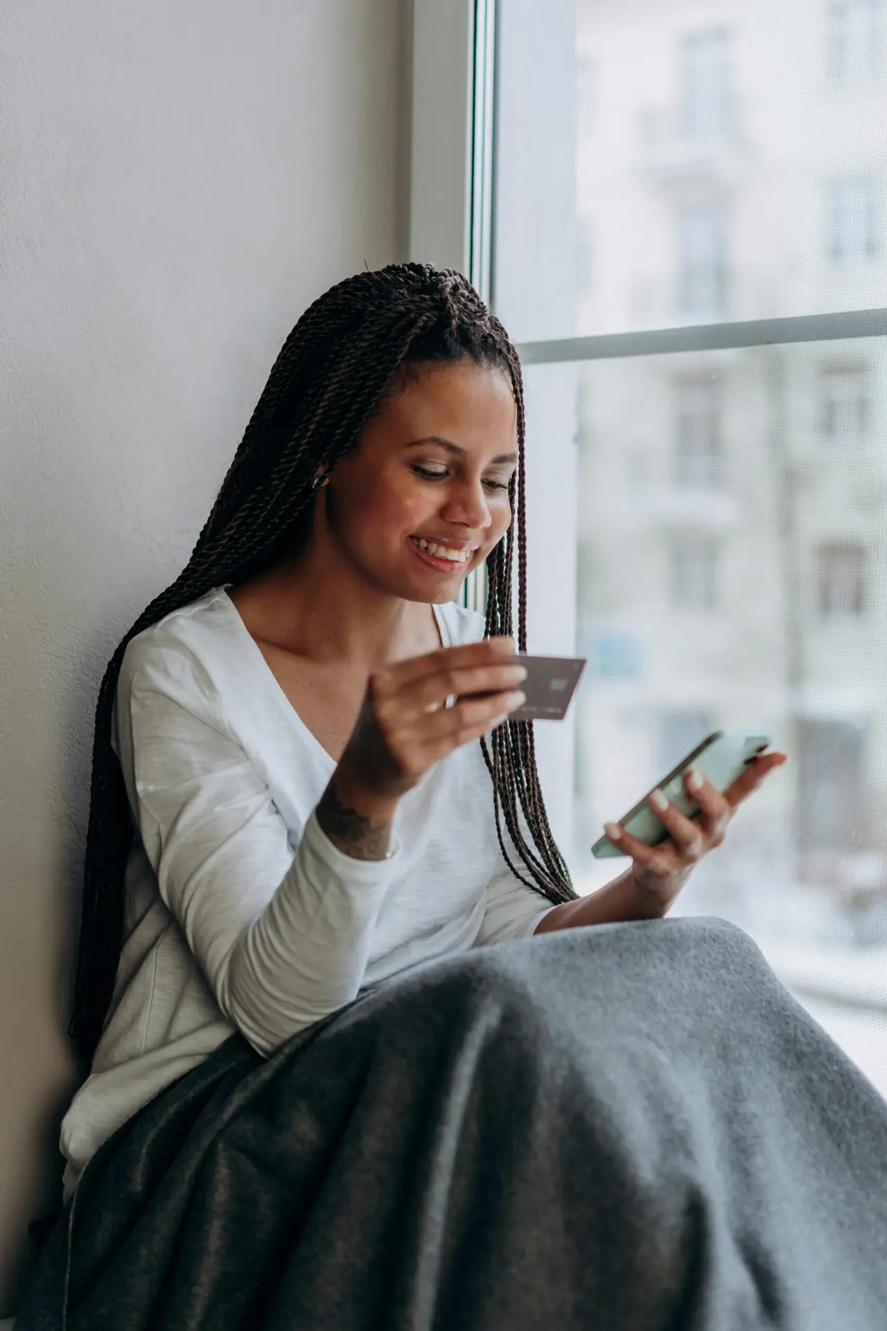 A smiling woman using a mobile phone and credit card for online shopping by the window.